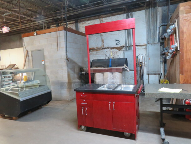 One Red Wood Work Station/Kiosk With A Black Laminate Top, 2 Full Size Hotel Pans, Push Bar,  4 Doors, And 2 Drawers, On Casters. 48X25X36